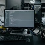 A cluttered workstation in an office featuring a monitor displaying code, surrounded by a keyboard, mouse, and wiring.